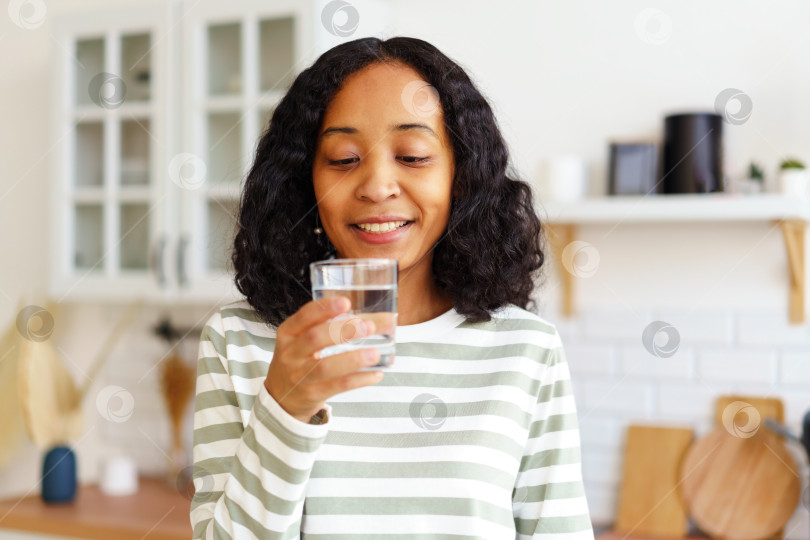 Скачать Улыбающаяся афроамериканка наслаждается стаканом чистой воды, стоя на кухне фотосток Ozero