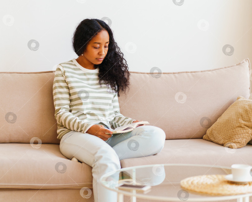 Скачать Красивая афроамериканка сидит на диване и читает книгу. Проводить время дома в одиночестве фотосток Ozero