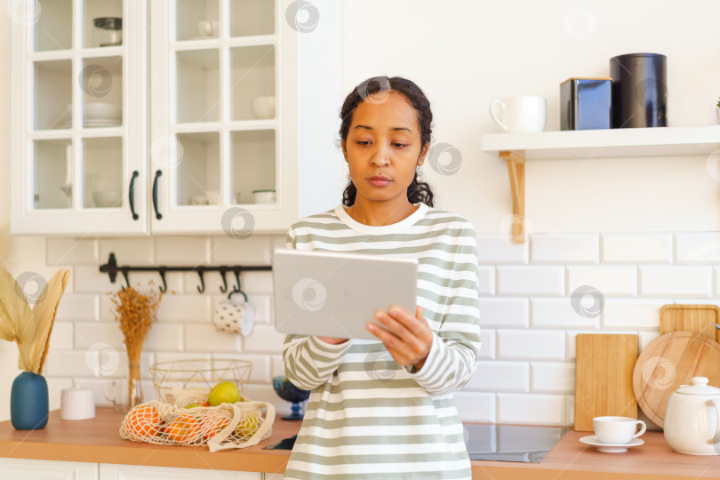 Скачать Афроамериканка ищет рецепт на планшетном компьютере перед приготовлением блюда на кухне фотосток Ozero