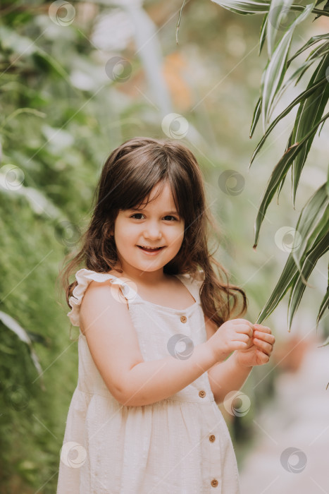 Скачать маленькая девочка брюнетка гуляет по ботаническому саду в хлопковом летнем платье. счастливый ребенок и пальмы фотосток Ozero
