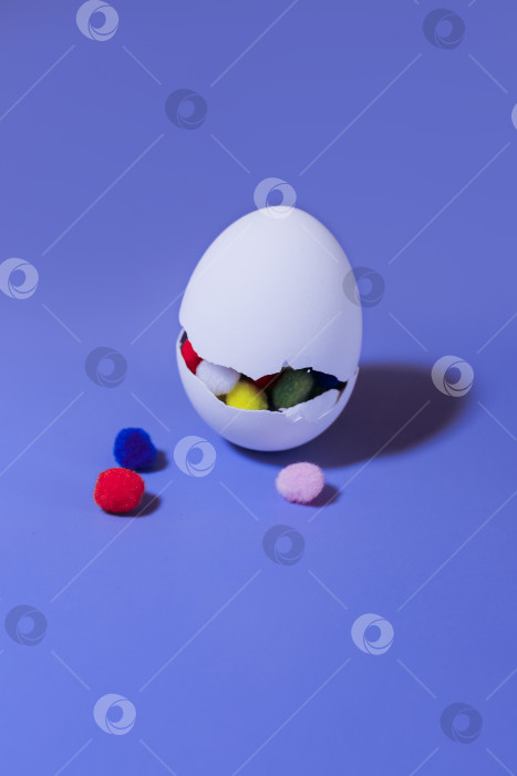 Скачать Белое яйцо с большой трещиной и цветными помпонами внутри на фиолетовом фоне фотосток Ozero