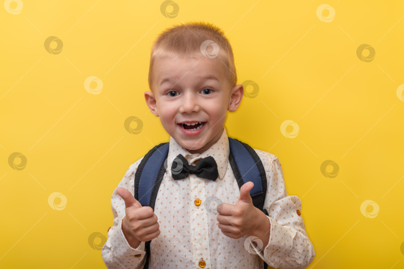 Скачать Обратно в школу. Белокурый забавный мальчик с рюкзаком в светлой рубашке на желтом фоне демонстрирует супер, класс и улыбается. Смотрит в камеру. Образование. фотосток Ozero