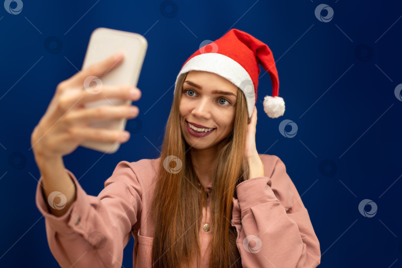 Скачать Девушка в шапке Санта-Клауса на изолированном синем фоне сфотографирована на свой телефон фотосток Ozero