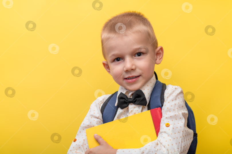 Скачать Обратно в школу. Белокурый забавный мальчик с рюкзаком в светлой рубашке на желтом фоне держит в руках книги и смотрит в камеру. фотосток Ozero