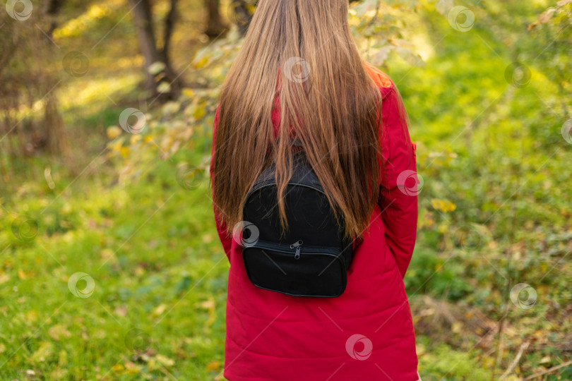 Скачать Молодая девушка в красной куртке с черным рюкзаком за спиной и длинными растрепанными волосами в лесу фотосток Ozero