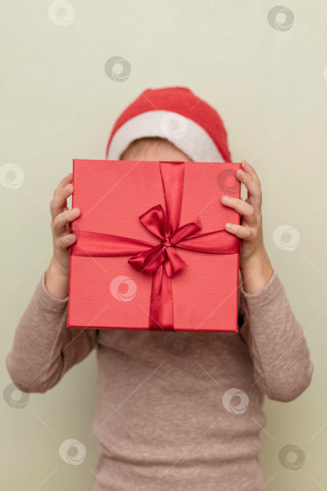 Скачать Забавный ребенок в шапке Санта-Клауса держит красную подарочную коробку, закрывая ею свое лицо. Концепция новогодних и рождественских подарков фотосток Ozero