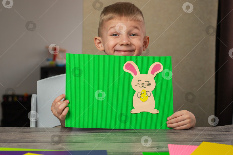 Скачать Радостный мальчик показывает открытку с кроликом, сделанную с помощью аппликации из разноцветной бумаги. Смастерьте пасхального кролика. фотосток Ozero