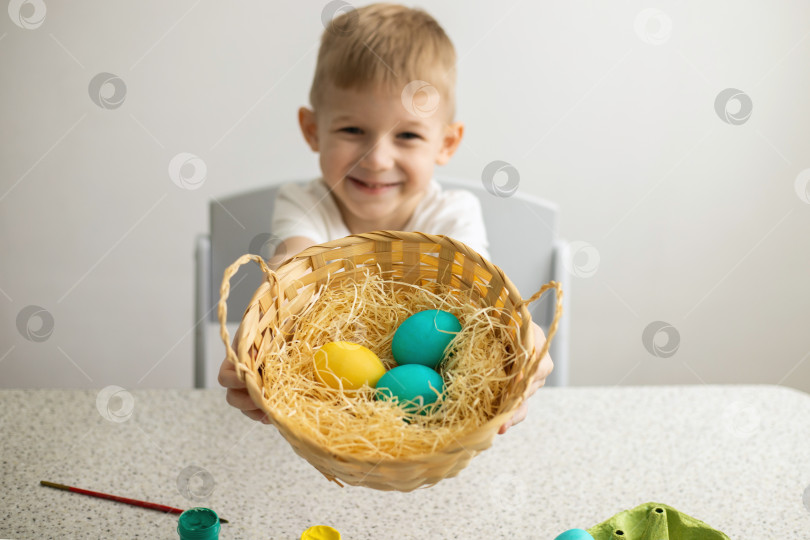 Скачать Счастливый ребенок показывает плетеную корзинку с крашеными яйцами. На столе лежат кисти и краски. Пасха. фотосток Ozero
