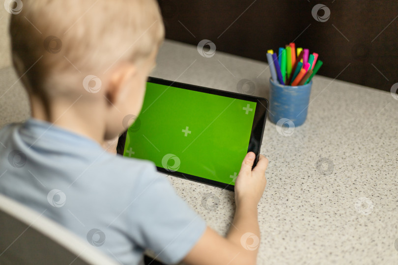 Скачать Ребенок сидит за столом, играет и смотрит на планшет на зеленом экране. Хромакей фотосток Ozero
