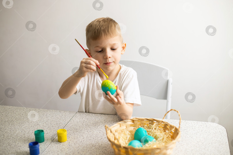 Скачать Ребенок, сидящий за столом, раскрашивает яйца кисточкой. Рядом лежат краски и плетеная корзина. Пасха фотосток Ozero