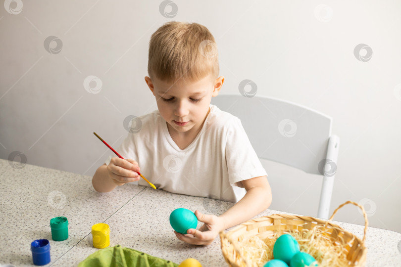 Скачать Ребенок, сидящий за столом, раскрашивает яйца кисточкой. Рядом лежат краски и плетеная корзина. Пасха фотосток Ozero