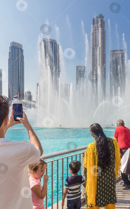 Скачать Дубай, Объединенные Арабские Эмираты - 20 декабря 2022 года: Люди-туристы смотрят на поющий танцующий фонтан возле Бурдж-Халифы. Достопримечательность Дубая. фотосток Ozero