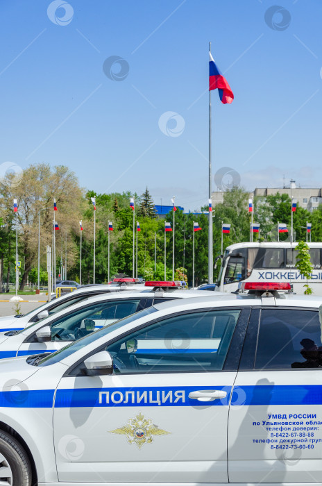 Скачать Ульяновск, Россия - 20 мая 2023 года: Дверь полицейской машины с логотипом и надписью POLICE и включенным проблесковым маячком на крыше. Красно-синяя мигалка полицейской машины. На заднем плане флаги России не в фокусе. фотосток Ozero