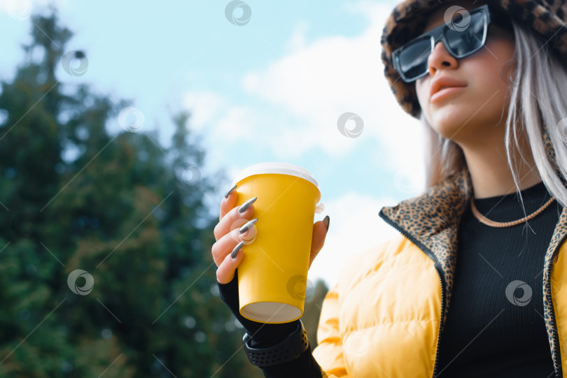 Скачать Молодая женщина, держащая в руках одноразовый стакан с напитком, на улице. Современная девушка в желтой куртке, очках и леопардовой шапке на прогулке. Сосредоточьтесь на чашке фотосток Ozero