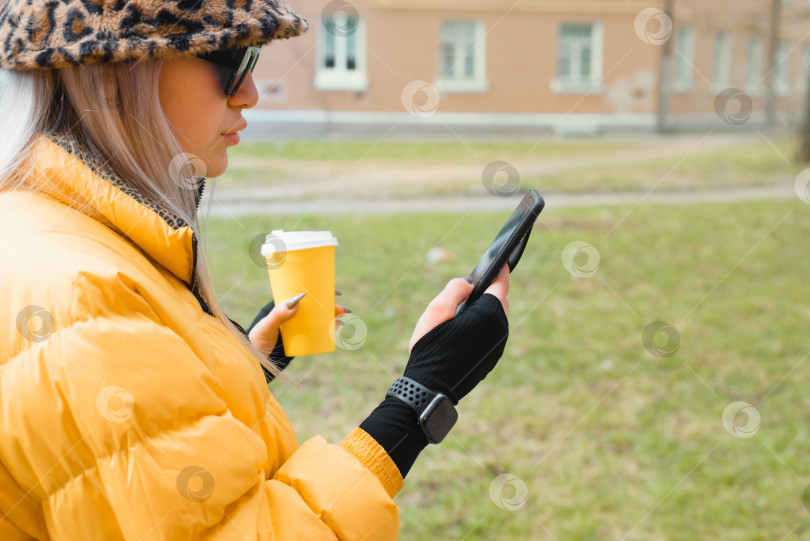 Скачать Молодая женщина болтает по телефону с чашкой кофе в руках на улице. Миллениал, хипстер в солнцезащитных очках, желтой куртке, леопардовой шляпе и умных часах. Выборочный фокус на руке фотосток Ozero