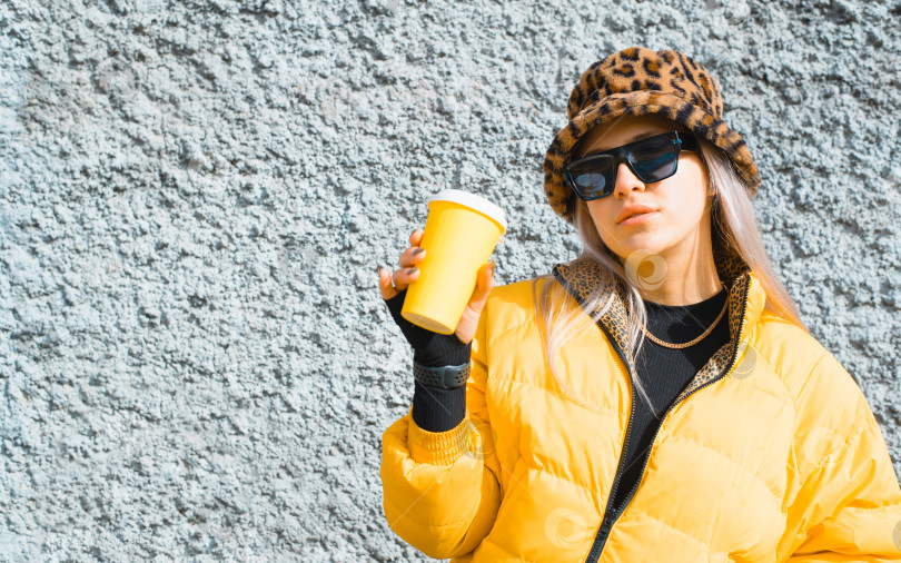 Скачать Молодая женщина в современной одежде, держащая одноразовый стаканчик с кофе, снаружи, на фоне стены. Портрет девушки в желтой куртке и очках. фотосток Ozero