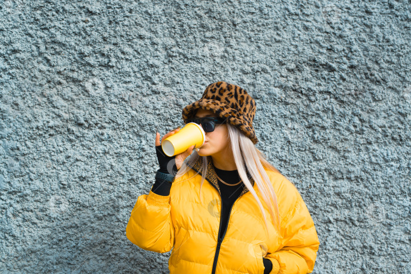 Скачать Женщина-хипстер пьет чашку кофе на улице на фоне стены. Миллениал в солнцезащитных очках, желтой куртке и леопардовой шляпе, расслабляющий образ жизни. фотосток Ozero