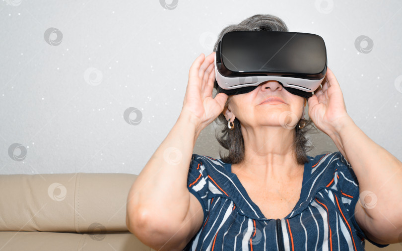 Скачать Пожилая женщина в очках виртуальной реальности. Пенсионерка, использующая очки виртуальной реальности в помещении, сидит на диване фотосток Ozero