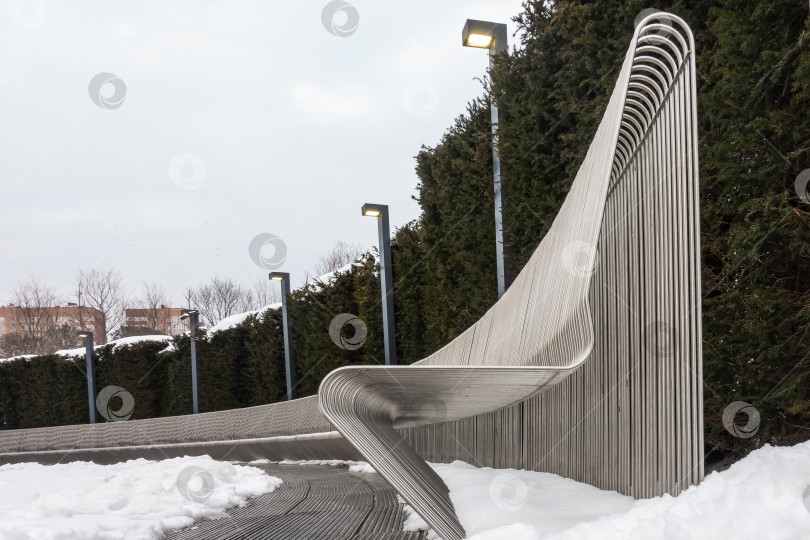 Скачать На заснеженной площади стоит стилизованная длинная изогнутая скамья, сделанная из металлических прутьев. фотосток Ozero