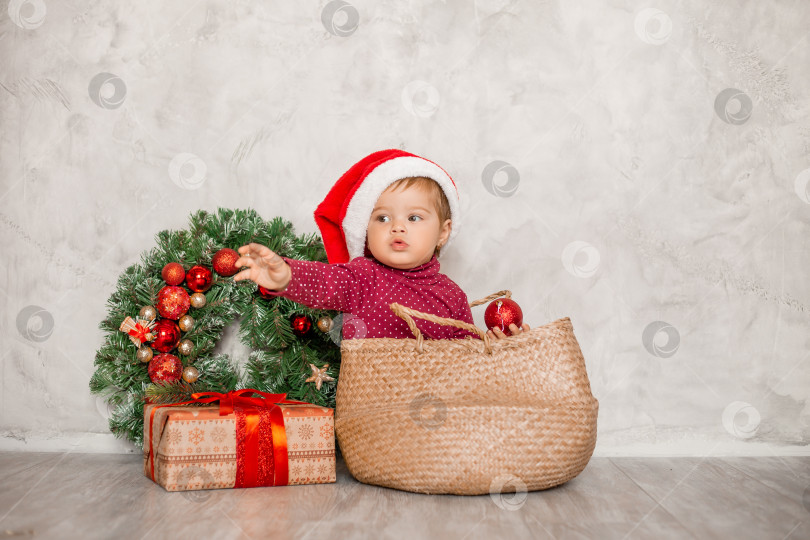 Скачать Милый малыш Санта сидит в плетеной корзине с подарочной коробкой и рождественской стеной на сером фоне. Рождественская концепция, текстовое пространство фотосток Ozero