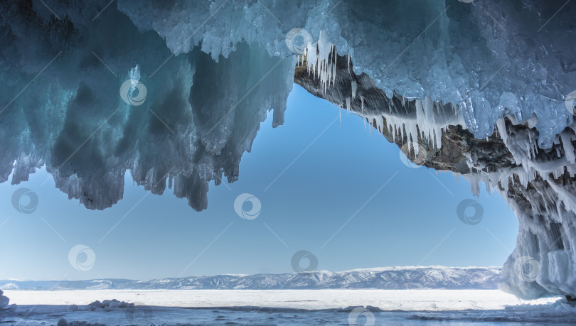 Скачать Покрытый льдом каменный грот. Причудливые ледяные сталактиты свисают с потолка на фоне лазурного неба фотосток Ozero