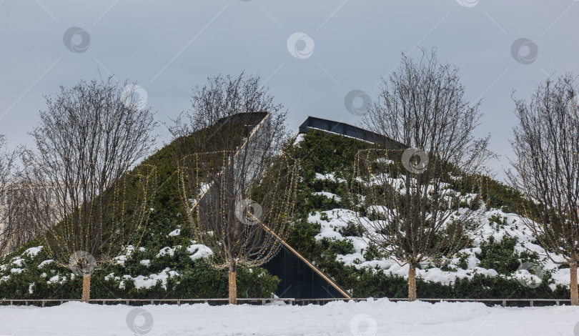 Скачать Декоративная горка в зимнем парке покрыта вечнозелеными кустарниками. фотосток Ozero