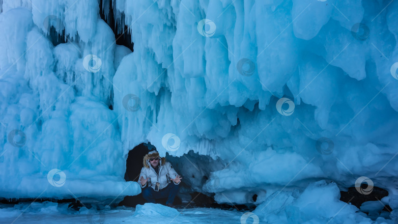 Скачать Скала полностью покрыта голубыми ледяными брызгами и сосульками. Мужчина в белом пуховике сидит в небольшом гроте. фотосток Ozero