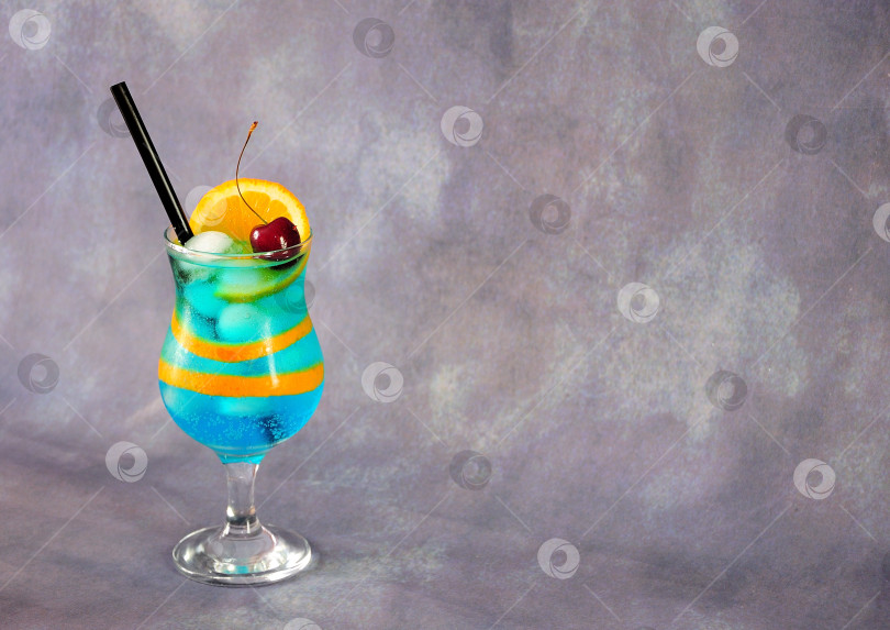 Скачать Высокий стеклянный бокал для коктейля "Голубая лагуна" с вишней и долькой апельсина на сером фоне. фотосток Ozero