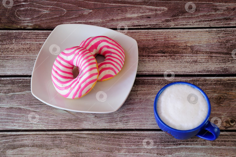 Скачать Чашка свежего капучино и два пончика в розовой глазури на деревянном столе. фотосток Ozero