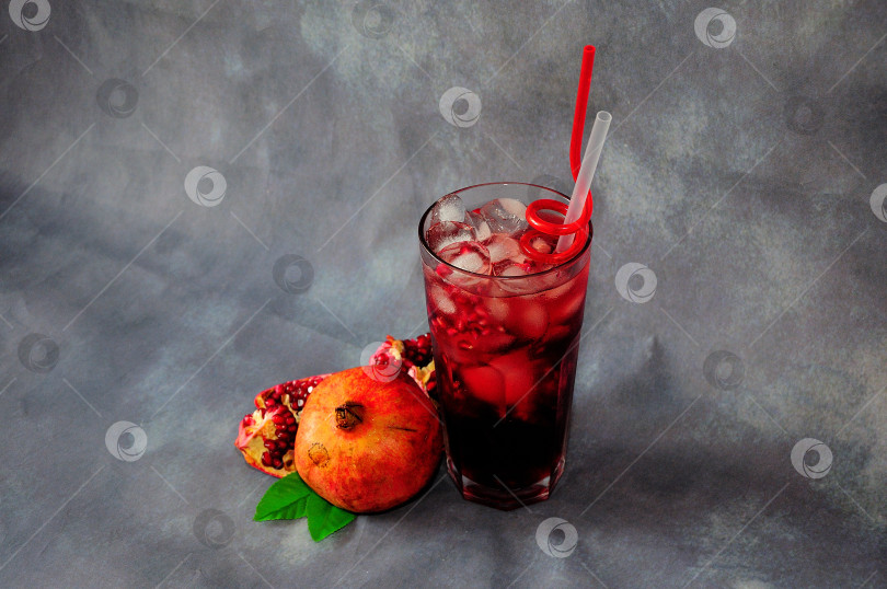 Скачать Стакан красного гранатового сока со льдом, соломинками и фруктовыми косточками рядом со спелыми фруктами на сером фоне. фотосток Ozero