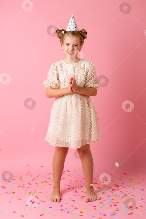 Скачать Счастливая маленькая девочка в праздничной шапочке сдувает со своих ладоней разноцветные конфетти на розовом фоне в студии фотосток Ozero