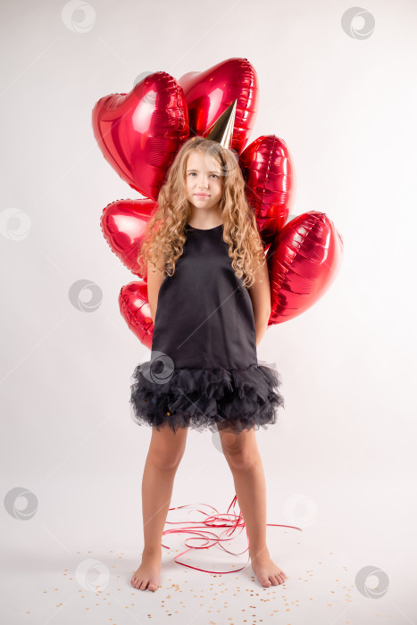 Скачать Счастливая маленькая девочка с охапкой шариков в виде сердечек и шапочкой на день рождения. конфетти на белом фоне. Пространство для текста фотосток Ozero