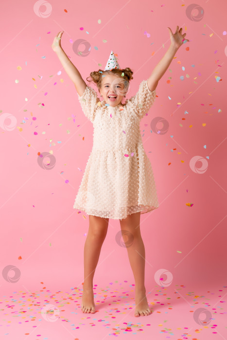 Скачать Счастливая маленькая девочка в праздничной шапочке сдувает со своих ладоней разноцветные конфетти на розовом фоне в студии фотосток Ozero