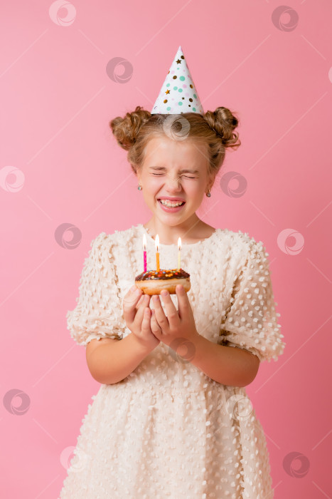 Скачать Счастливая маленькая девочка в шапочке на свой день рождения загадывает желание и задувает свечи на торте. Розовый фон, текстовое пространство фотосток Ozero
