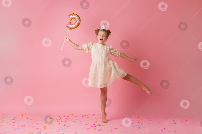 Скачать счастливая маленькая девочка в праздничной шапочке лежит на розовом фоне с конфетти, держит руки у лица. Пространство для текста фотосток Ozero