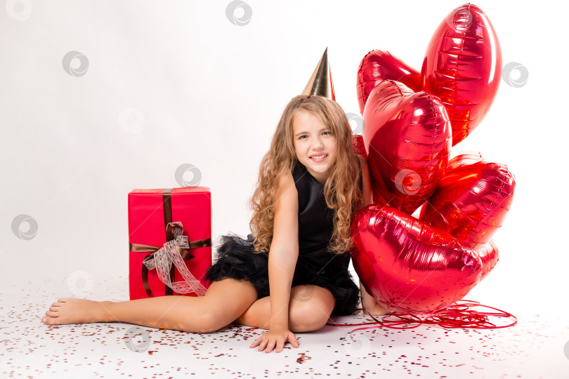 Скачать Счастливая маленькая девочка с охапкой шариков в виде сердечек и шапочкой на день рождения. конфетти на белом фоне. Пространство для текста фотосток Ozero
