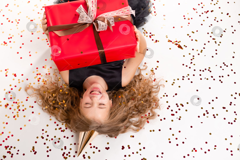 Скачать Счастливая маленькая девочка лежит на белом фоне с подарочной коробкой и шапочкой на свой день рождения. Вид сверху, место для текста фотосток Ozero