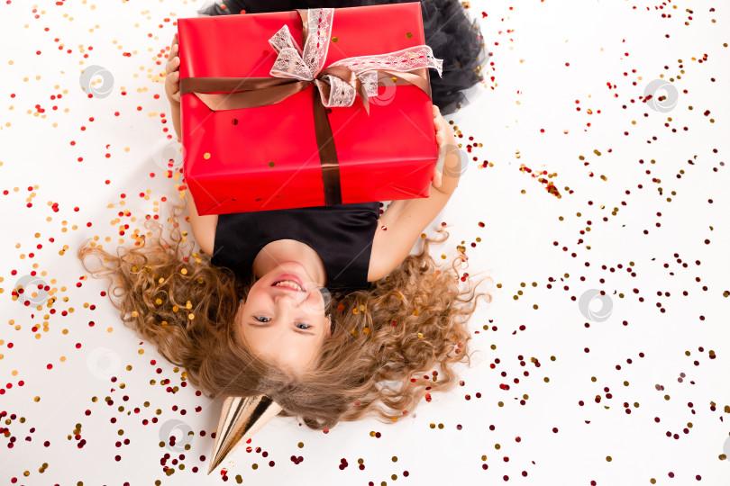 Скачать Счастливая маленькая девочка лежит на белом фоне с подарочной коробкой и шапочкой на свой день рождения. Вид сверху, место для текста фотосток Ozero