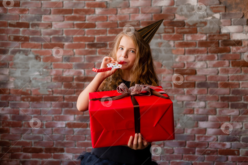 Скачать счастливая маленькая девочка в праздничной шапочке держит в руках большую красную подарочную коробку. кирпичная стена. концепция праздника, место для текста фотосток Ozero