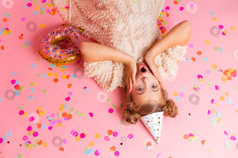 Скачать счастливая маленькая девочка в праздничной шапочке лежит среди разноцветных конфетти на розовом фоне, вид сверху. Пространство для текста фотосток Ozero