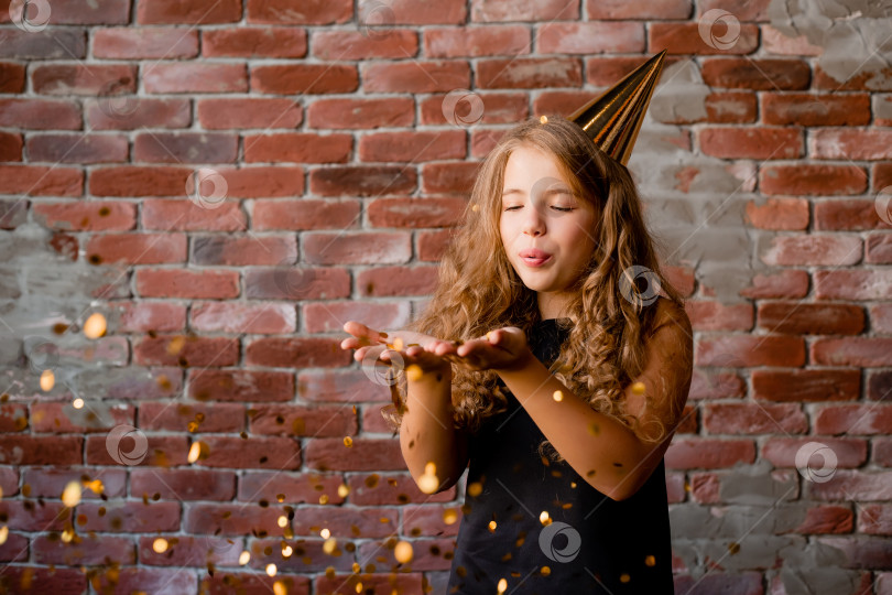 Скачать Счастливая маленькая девочка в шапочке на свой день рождения сдувает с ладоней конфетти. концепция праздника, кирпичный фон, текстовое пространство фотосток Ozero