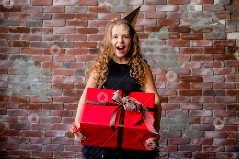 Скачать счастливая маленькая девочка в праздничной шапочке держит в руках большую красную подарочную коробку. кирпичная стена. концепция праздника, место для текста фотосток Ozero