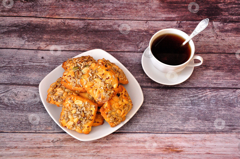 Скачать Тарелка с горкой песочного печенья с семечками и орехами на деревянном столе, рядом с чашкой горячего черного кофе. фотосток Ozero