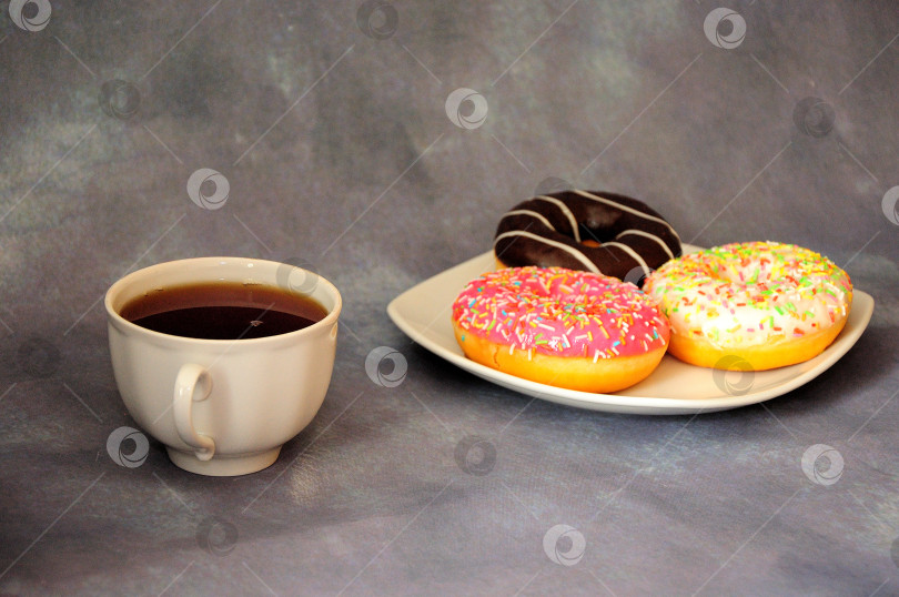Скачать Чашка черного чая и тарелка с тремя глазированными пончиками на сером фоне. фотосток Ozero