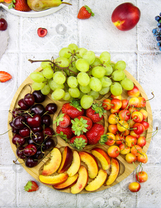 Скачать Богатая фруктовая тарелка, наполненная фруктами и ягодами. Ассорти из черной вишни, зеленого винограда, желтой вишни, персиков и клубники на деревянной тарелке на столе. Ассорти из сырых фруктов и ягод фотосток Ozero
