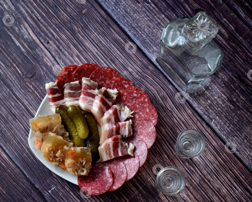 Скачать Бутылка и два стакана водки стоят на деревянном столе рядом с тарелкой с разнообразными мясными закусками. фотосток Ozero