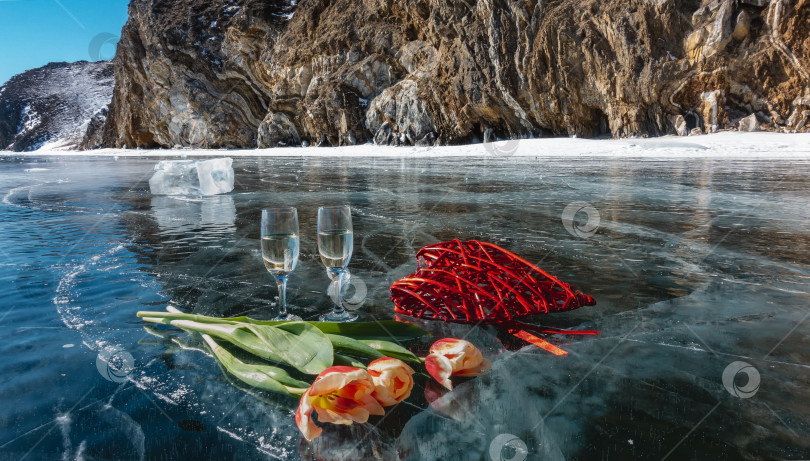 Скачать На замерзшем озере стоят два бокала с шампанским. Рядом лежат красные и желтые тюльпаны, декоративное плетеное сердечко, осколок льда. фотосток Ozero