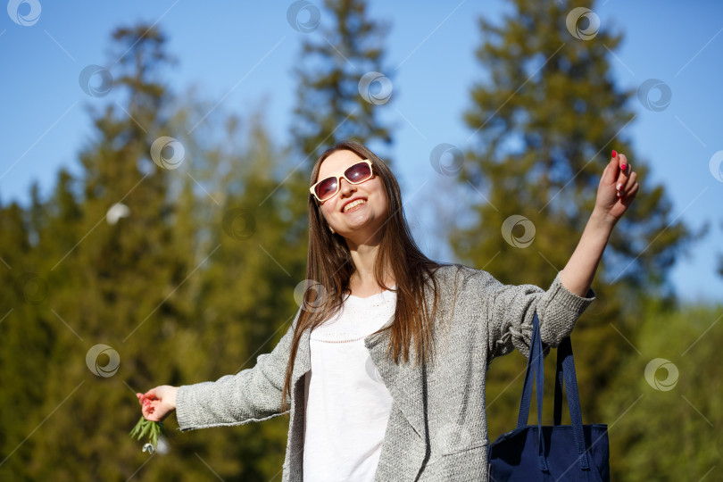 Скачать Счастливая женщина в парке в солнцезащитных очках, улыбающаяся с поднятыми руками. Сумка в руке. фотосток Ozero