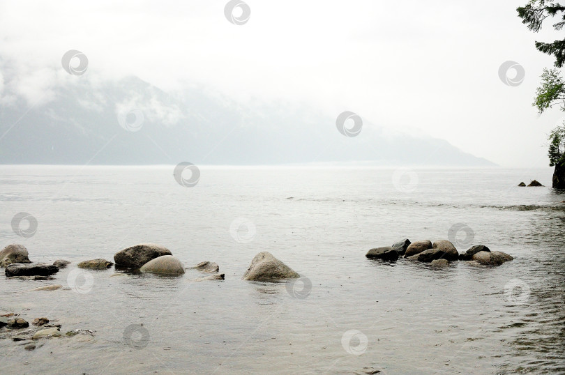 Скачать Ряд больших камней, торчащих из озера, окружен высокими горами, над которыми идет дождь. фотосток Ozero