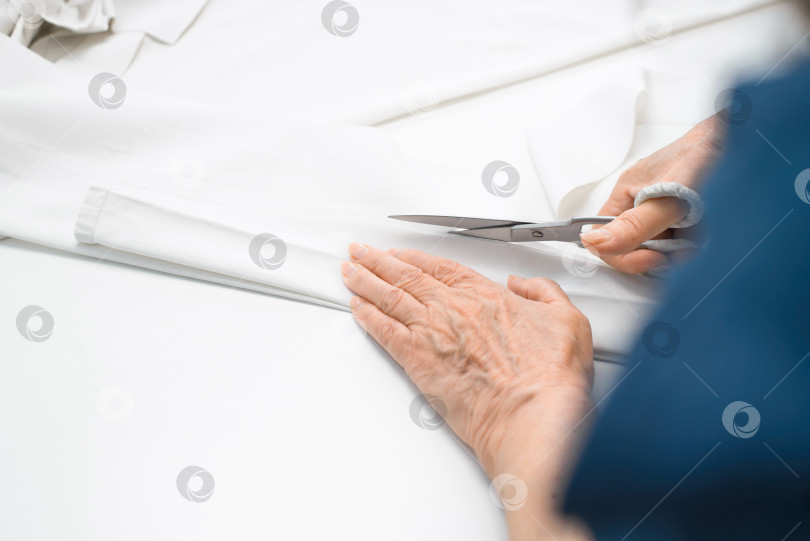 Скачать Пожилая женщина режет ножницами белую ткань, руки крупным планом. Старшая женщина-портниха. Работа на пенсии или хобби дома. фотосток Ozero